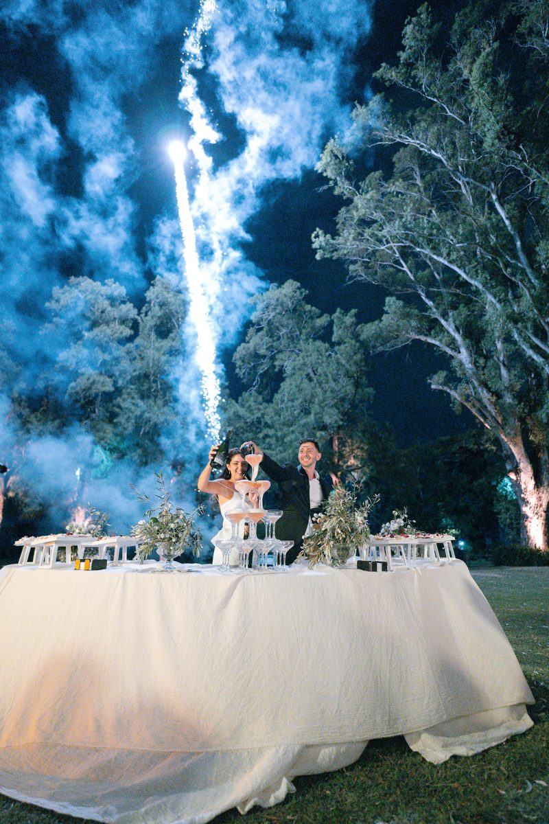 Los novios brindando con una torre de copas y una fuente de chispas frías detrás, de noche en el parque