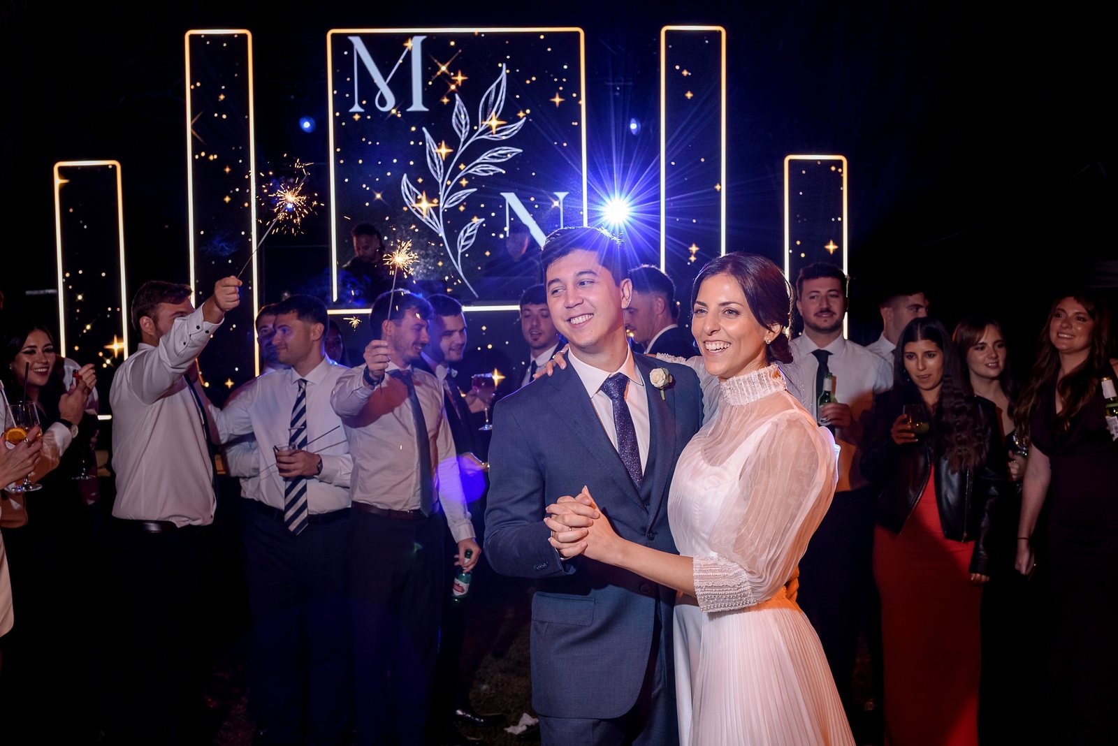 Los novios en su primer baile rodeados de invitados con chispas y el cartel iluminado M N